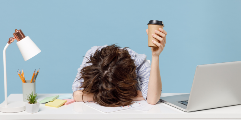 Woman holding coffee cup has her head down on her desk between a laptop and desk lamp