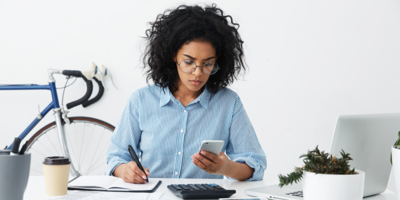 Woman in a home office concentrating on taking notes while looking at her cell phone.