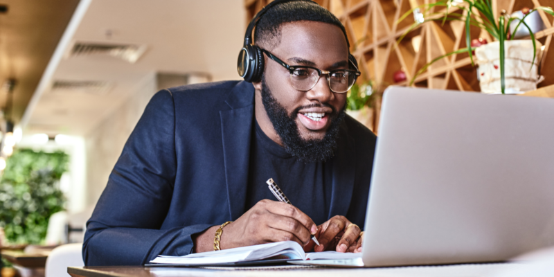 Man wearing headphones looking excited as he takes notes while looking at his laptop.
