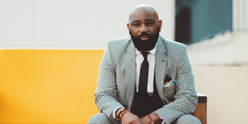 A serious adult bearded bald African man in an elegant plaid suit with a tie is sitting on a yellow soft sofa outdoors and looking at the camera with a determined expression on his face