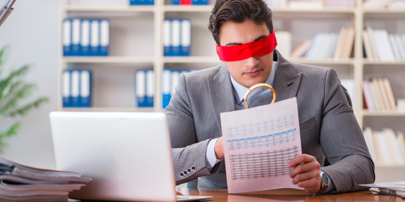 Blindfold businessman sitting at desk in office with a magnifying glass and a sheet of paper
