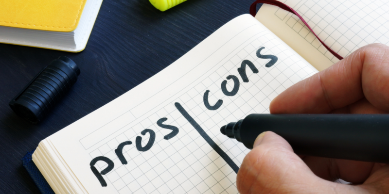 Hand holding a black marker hovers over an open journal with grid-lined paper and the word "pros" on the left and "cons" on the right