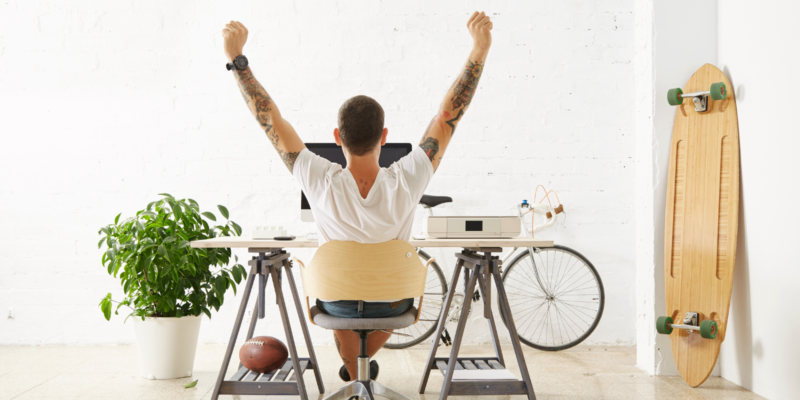 Man sitting in a while office lifting his hands up in joy. There is a bicycle and skateboard in the background and he has an industrial style desk.