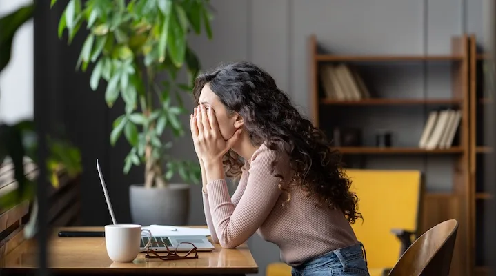 Tired overworked female employee covering face with hands and thinking about solution while sitting at table with laptop