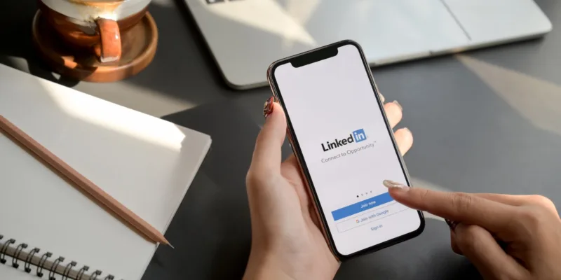 Person touching iPhone with LinkedIn screen with laptop and notebook on desk in the background