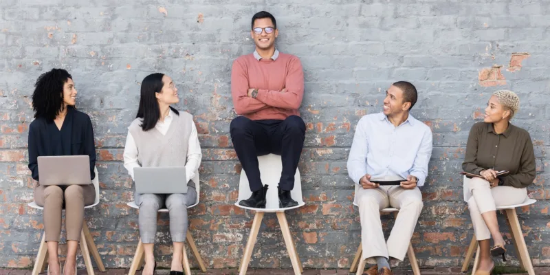 5 people sitting in chairs in a line against a brick wall for interview, meeting or opportunity and the man in the middle standing out from the others.