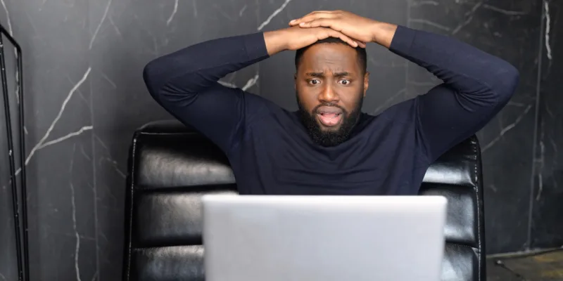 Image of shocked black businessman holding hands on head, looking very disappointed, looking at screen of computer eyes wide open,