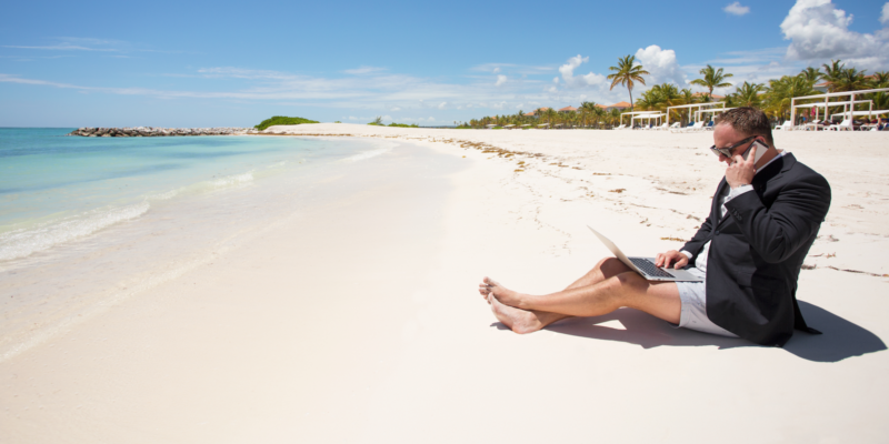 Man in suit jacket sitting on the beach with bathing suit on working on laptop and on the phone