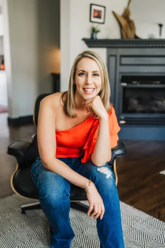 Nicki Krawczyk, founder of Fired Up Freelance, sits in a black swivel chair in an orange top and jeans with her head resting on her fist, which rests on her leg.