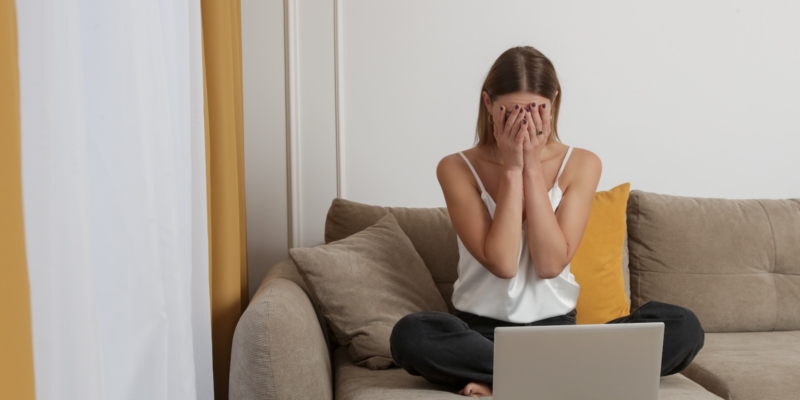 Woman sitting on the couch with her laptop with her hands in her head worried about news she's received.